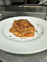 A slice of baked lasagna with layers of cheese and sauce is served on a white plate, garnished with chopped herbs. The background shows a stainless steel kitchen setting.