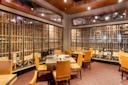 Private dining room with tan leather chairs around tables, place settings ready, and large glass windows displaying an extensive wine collection in wooden racks along two walls. Warm lighting and red-patterned carpet.
