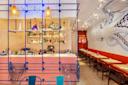 A vibrant bar with a nautical theme features a colorful counter, a variety of bottles on shelves, and blue wire decor. Red seating lines the right wall adorned with a large octopus mural. Bright lights and a neon sign create a lively atmosphere.