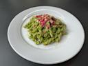 A plate of twisted pasta coated in green pesto sauce, topped with small cubes of red and white cured meat, served on a white plate against a dark background.