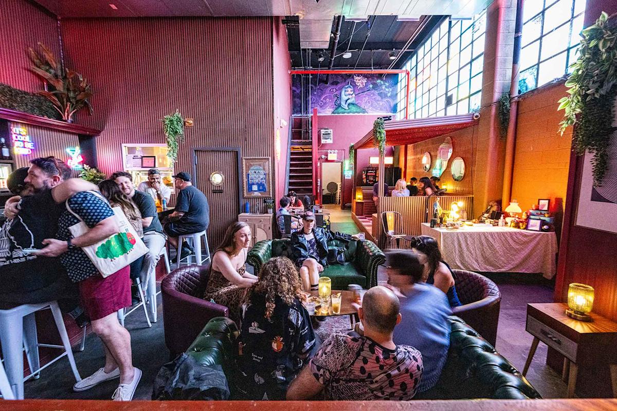 A lively bar with people socializing on couches and bar stools. Colorful lights, large windows, plants, and eclectic decor create a vibrant, cozy atmosphere. A mural is visible above a red staircase in the background.