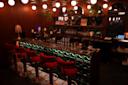 A dimly lit bar with a modern, cozy interior. Red velvet bar stools line a marble counter illuminated from below. Plants hang from the ceiling, and round pendant lights add warmth to the space. Shelves with glasses and bottles are in the background.