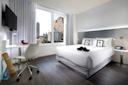 Modern hotel room with a large bed, white bedding, and decorative pillows. A flat-screen TV is mounted on the wall. There's a desk with a chair, a yoga mat, and a large window revealing a city skyline. Soft lighting creates a cozy atmosphere.