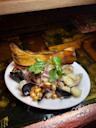 A plate of seafood ceviche with clams, mussels, hominy corn, toasted corn, red onions, cilantro, and two large plantain chips, served on a white dish on a wooden counter.