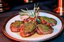 A plate of grilled lamb chops topped with a vibrant green herb sauce, garnished with sprigs of rosemary. The chops are arranged on a round, white plate with a dark rim, set against a dark background.