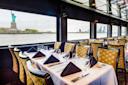 Elegant dining tables set with black napkins and glassware on a boat, with large windows offering a view of the Statue of Liberty and the New York City skyline across the water.