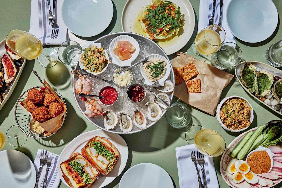 A table set with oysters, shrimp, spreads, crackers, sandwiches, falafel, salad, and vegetables, surrounded by plates, napkins, and glasses of white wine.