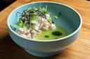 A light blue bowl containing diced raw fish dressed in a green sauce, garnished with thin black strips and microgreens, sits on a wooden table. The dish is artfully presented, highlighting its fresh ingredients.
