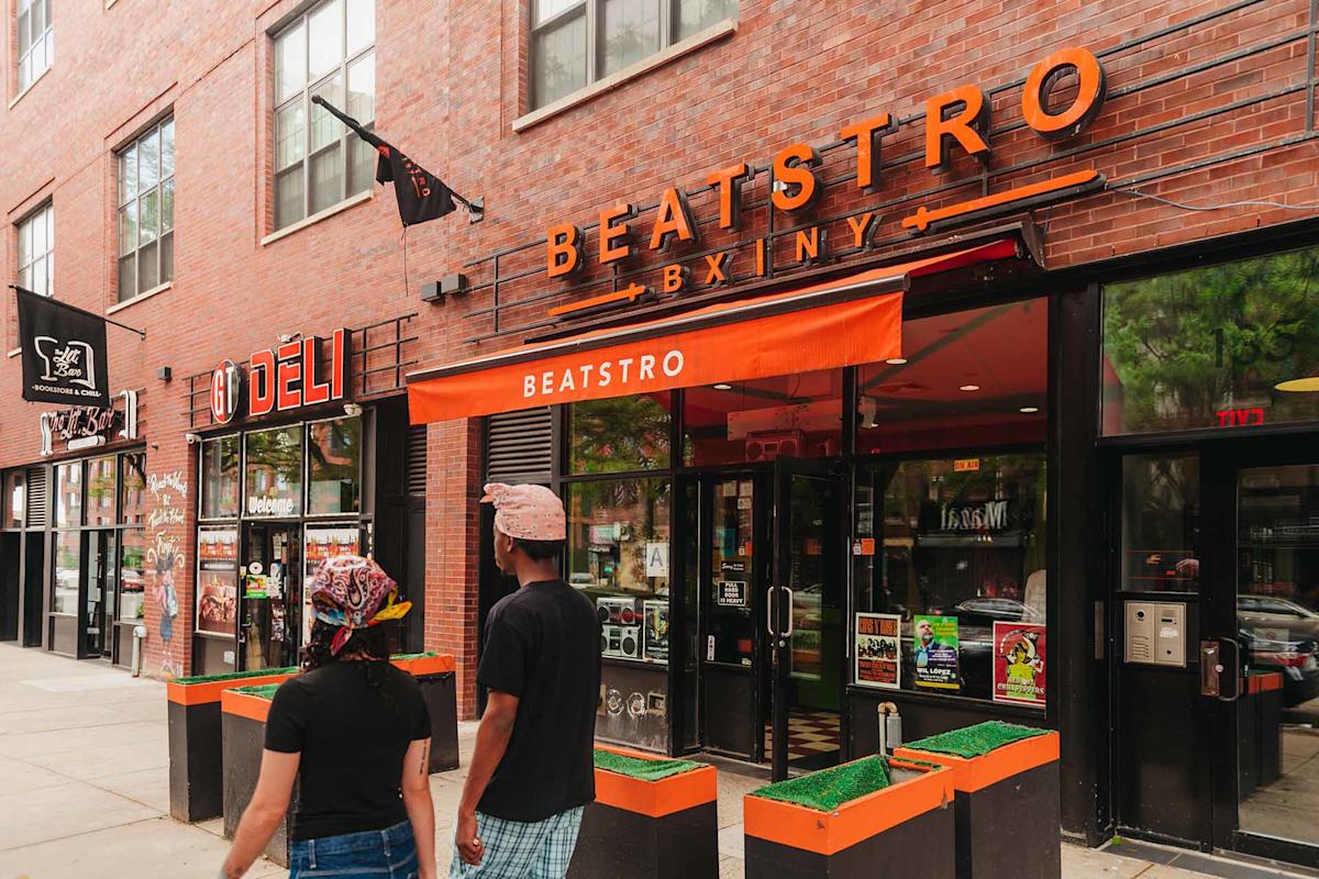 View from an elevated subway platform looking down onto a city street with crosswalks, pedestrians, trees, parked cars, food trucks, and buildings with graffiti under a clear sky.