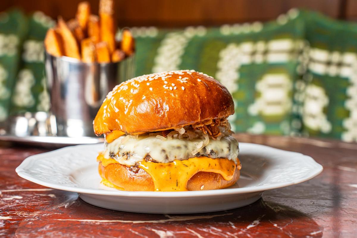 A cheeseburger with melted cheese and crispy onions on a sesame bun sits on a white plate, with a metal cup of fries in the background.