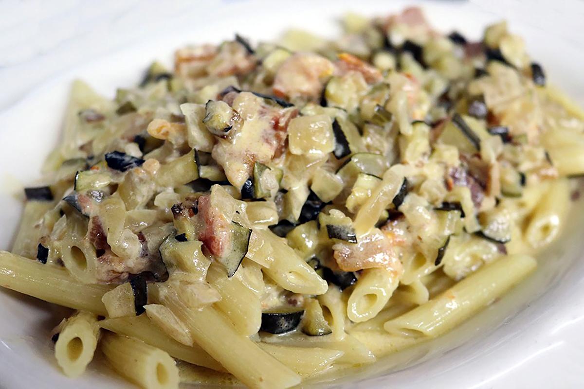 A plate of penne pasta topped with a creamy sauce containing diced zucchini, onions, and bits of bacon or ham. The sauce appears rich and thick, coating the pasta evenly.