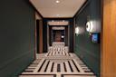 A modern hallway with dark green walls, geometric black and white patterned carpet, circular wall lights, and a wall-mounted screen, leading to a distant door.