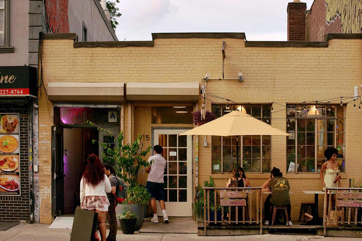 People relax and dine at outdoor tables under a yellow umbrella in front of a small brick restaurant. Plants decorate the patio, and a person opens the front door. The scene appears calm and inviting.