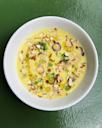 A white bowl filled with yellow soup, garnished with chopped almonds, sliced green onions, and herbs, sits on a textured green surface.