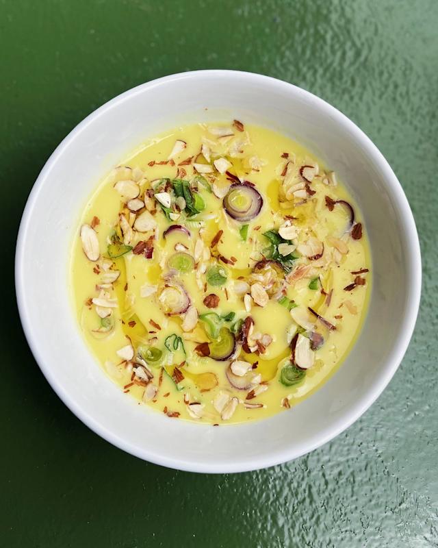 A white bowl filled with yellow soup, garnished with chopped almonds, sliced green onions, and herbs, sits on a textured green surface.