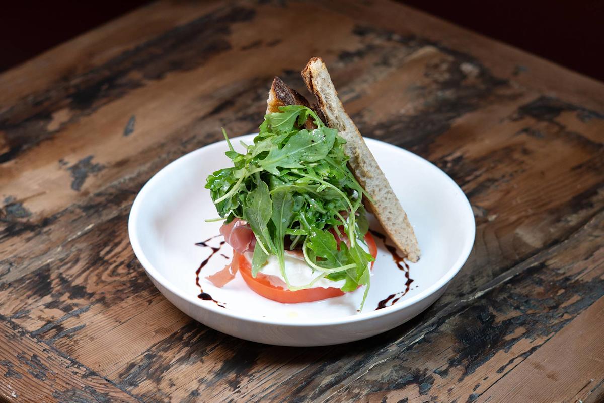 A white plate on a rustic wooden table holds a salad with arugula, tomato, mozzarella, and prosciutto, drizzled with balsamic glaze, and served with a slice of toasted bread.
