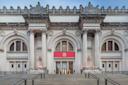 Facade of a grand museum with neoclassical architecture featuring large columns, arched windows, and ornate detailing. A red banner with "The Met" hangs above the entrance. Stone steps lead up to the entrance.