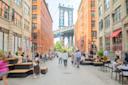 A busy cobblestone street in Brooklyn, New York, with people sitting and walking, tall brick buildings on both sides, and the Manhattan Bridge visible in the background.