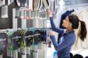 A woman wearing a blue jacket selects a pickleball paddle from a wall display in a sports store. Several paddles are arranged in rows, and clothing items hang on an adjacent rack.