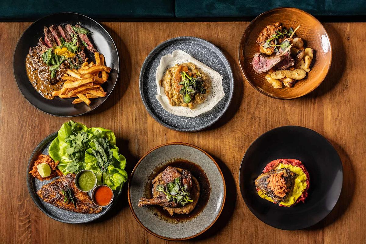 Six plates of assorted gourmet dishes, including steak with fries, grilled chicken, roasted meat with potatoes, a vegetable-topped dip, and dishes with colorful sauces, all arranged on a wooden table.
