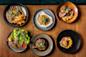 Six plates of assorted gourmet dishes, including steak with fries, grilled chicken, roasted meat with potatoes, a vegetable-topped dip, and dishes with colorful sauces, all arranged on a wooden table.