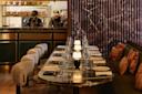 A sophisticated restaurant interior with a marble dining table set for eight, cushioned chairs, decorative pillows, dim lighting, and two chefs working behind a counter in the background.