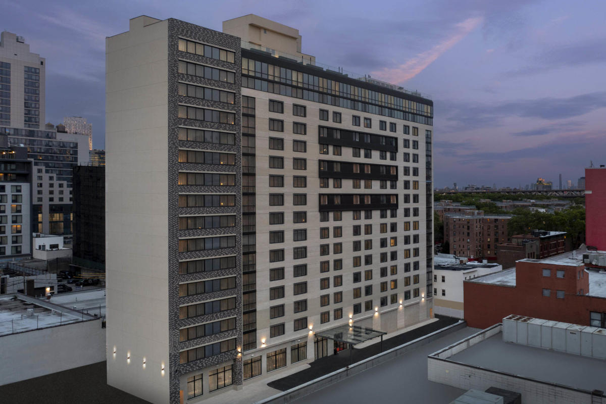 A tall, modern apartment building at dusk, with numerous windows illuminated. The building features a mix of light and dark panels and stands amidst an urban landscape, under a clouded sky.