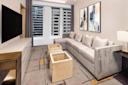 A modern living room features a gray L-shaped sofa, abstract art on the wall, a large flat-screen TV, and two square wooden coffee tables. Large windows reveal a cityscape of buildings. The decor is neutral with contemporary accents.