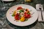 A plate of heirloom tomato salad with sliced red and yellow tomatoes, burrata cheese, microgreens, and a drizzle of balsamic glaze. A fork and knife are placed on a folded napkin to the side on a marble table.