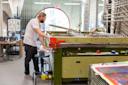 A person with long hair and a beard stands at a large green screen printing machine in a workshop, focusing on their work. The room is filled with equipment, stacks of materials, and tools scattered around.