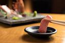 A piece of sashimi being held with chopsticks is dipped into soy sauce, with a plate of assorted sashimi and garnishes blurred in the background on a wooden table.