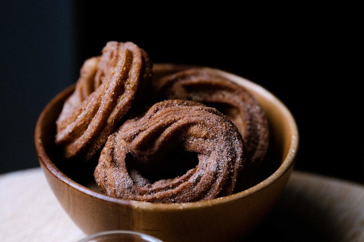 A wooden bowl filled with three round, ridged, sugar-coated churros sits on a light surface, with a dark background behind them.