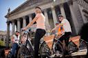A group of smiling people pause on their rental bikes in front of a grand, columned building. The bikes display “Holland Bikes” signs. One woman looks back at the camera, and the group appears ready to ride.