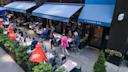 A busy outdoor restaurant scene on a city sidewalk, with people dining at tables under blue awnings and red Aperol Spritz umbrellas. The entrance reads "41 Madison Ave." Green plants decorate the area.