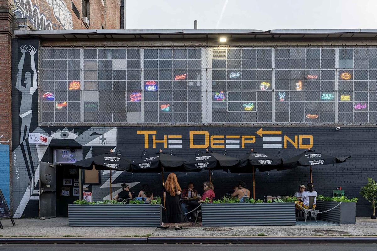 People sit under black umbrellas at outdoor tables in front of “The Deep End” bar, with a mural and neon signs on the building’s upper windows.