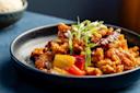 A plate of stir-fried chicken with bell peppers, onions, and a garnish of green onions is served on a dark round plate. A whole red chili pepper adds a hint of spiciness. A bowl of rice is blurred in the background.