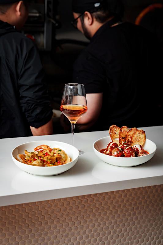Two bowls of food and a glass of rosé wine on a white counter; one bowl contains shrimp and vegetables, the other has meatballs with toasted bread. Two people are working in the background.