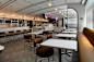 A modern café with brown chairs, white tables, and a counter displaying pastries. Signs above the counter read "Egg Creams" and "Russ & Daughters." Hanging globe lights illuminate the sleek, minimalist design.