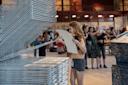 A person is lifting a sheet of paper from a large drying rack in an art studio. Other individuals are seen in the background, some taking photos. The space is well-lit and features high ceilings and exposed beams.