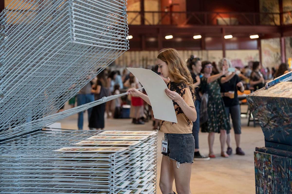 A person is lifting a sheet of paper from a large drying rack in an art studio. Other individuals are seen in the background, some taking photos. The space is well-lit and features high ceilings and exposed beams.