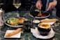 A table set with a green marble surface holding two plates of food, including a small salad and a dish topped with melted cheese. There are breadsticks, glasses of red and white wine, and a pepper grinder.