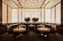 Elegant, modern restaurant dining room with round tables set for guests, leather chairs, warm lighting, tall windows with sheer curtains, and two large potted plants as a central feature.