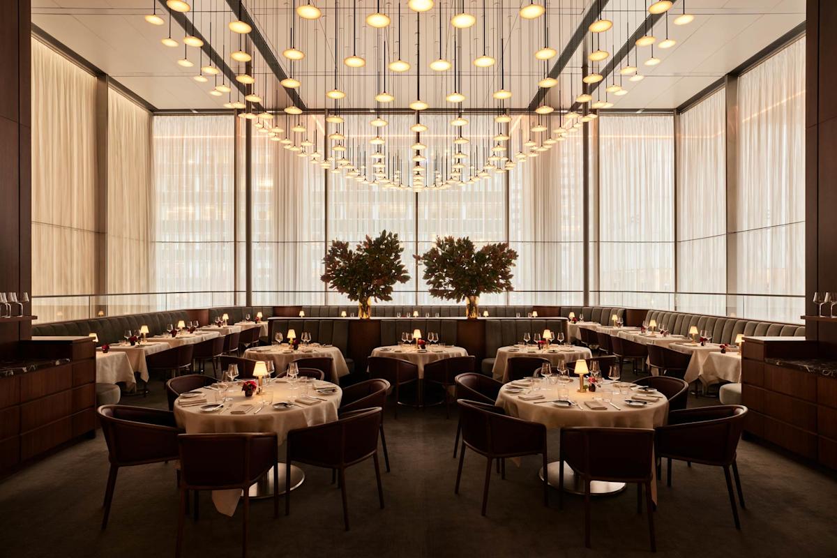 Elegant, modern restaurant dining room with round tables set for guests, leather chairs, warm lighting, tall windows with sheer curtains, and two large potted plants as a central feature.