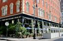 Street view of a red-brick corner building with arched windows, a black storefront labeled "Roman and Williams Guild NY," outdoor dining area with blue umbrellas, greenery, and street signs on a sunny day.