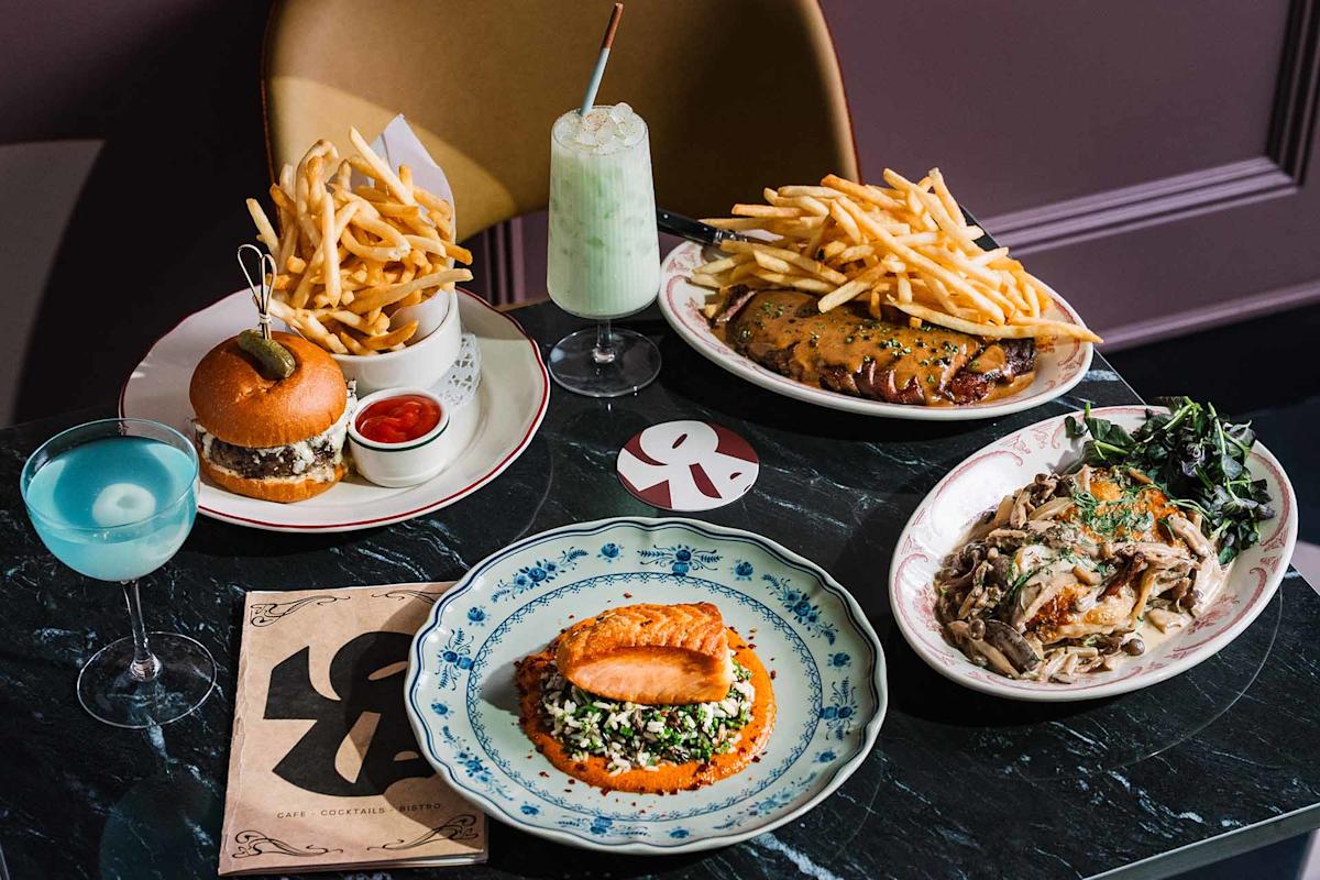 A table set with a burger and fries, ribs with fries, pasta, grilled salmon with vegetables, a blue cocktail, a green milkshake, and a menu on a dark marble surface.