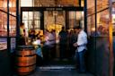 A group of people gather inside an open brick building labeled "Widow Jane," with a wooden barrel by the entrance and a man standing outside. Warm lighting glows from inside, creating a lively, welcoming atmosphere.