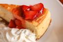 A slice of cheesecake topped with glazed strawberries and served with a dollop of whipped cream on a white plate.