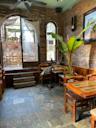 Cozy cafe with brick walls, wooden tables and chairs, a large leafy plant, and sunlight streaming through large windows and a glass door leading to an outdoor patio area.