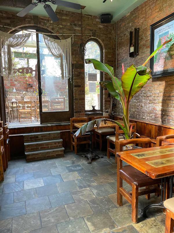 Cozy cafe with brick walls, wooden tables and chairs, a large leafy plant, and sunlight streaming through large windows and a glass door leading to an outdoor patio area.
