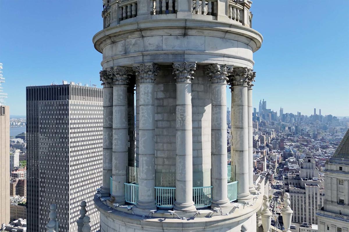 Close-up of a classical stone tower with columns and ornate capitals, overlooking a cityscape with tall buildings and a clear blue sky in the background.
